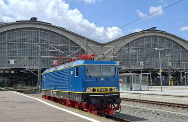 einzelne blaue Elektrolok BR243, im Hintergrund ist ein modernes Bahnhofsgebäude zu sehen