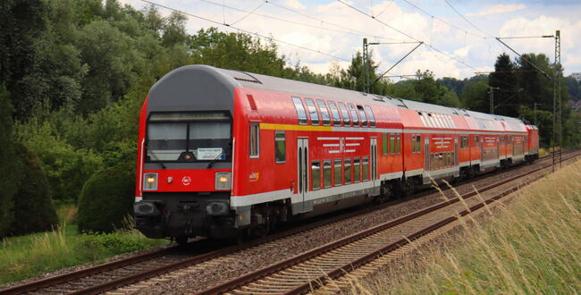 Roter ÖPNV Doppelstockzug fährt auf Gleisen durch grüne Landschaft mit Bäumen und Stromleitungen.