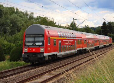 Roter ÖPNV Doppelstockzug fährt durch grüne Landschaft mit Bäumen und Stromleitungen.